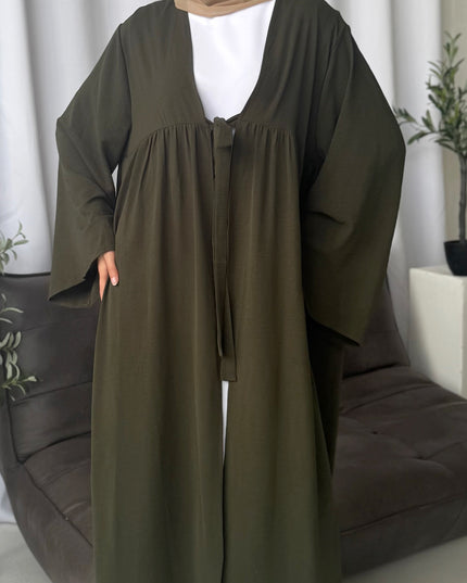 Woman wearing an olive green abaya with a white top, standing against a neutral background.