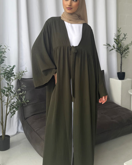 Woman wearing a long olive green dress with a matching hijab in a room with white curtains and plants.