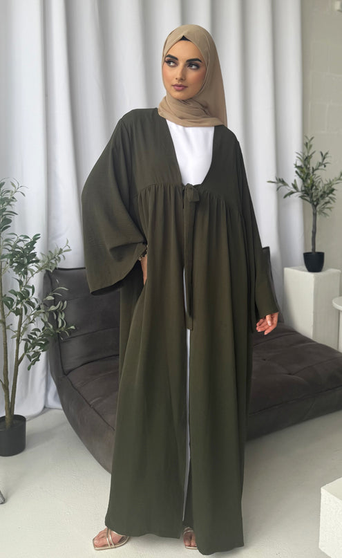 Woman wearing a long olive green dress with a matching hijab in a room with white curtains and plants.